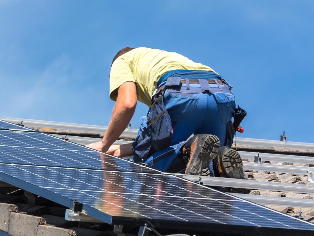 Instalador de costas, ajoelhado no telhado, encaixando uma placa solar nos trilhos sob céu azul.